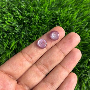 May include: Two round, polished gemstones with a translucent, purplish-pink hue. The stones are set against a backdrop of green artificial grass. The gemstones are small, likely for jewelry.