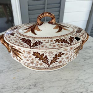 May include: An antique lidded serving dish with a floral and leaf pattern in brown and gold on a cream background. The dish has a handle on the lid and two side handles. The design is repeated on the lid and the body of the dish.