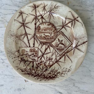 May include: A vintage ceramic plate with a brown bamboo and landscape design. The plate features three framed scenes: a deer scene, a house, and a bird scene. The plate has a cream-colored background.