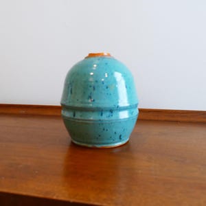 Pode incluir: Um vaso de cerâmica turquesa com corpo arredondado e borda laranja. O vaso tem uma textura salpicada e um design de dois níveis. Está sobre uma superfície de madeira.