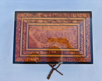 May include: Rectangular wooden jewelry box with a glossy finish. The box features a central panel with a unique wood grain pattern, framed by concentric borders of varying wood tones and a black border. A decorative tassel hangs from the front.