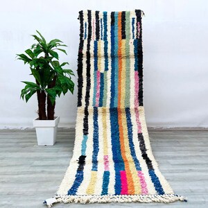 Puede incluir: Una alfombra larga y estrecha con rayas de colores sobre un fondo blanco. La alfombra está hecha de lana y tiene flecos en ambos extremos. La alfombra es de varios colores, incluyendo azul, rosa, amarillo y naranja.