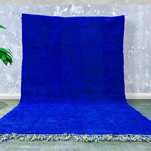 Op de afbeelding: Een levendig blauw tapijt met een zachte textuur, rechthoekig van vorm. Het tapijt heeft een crème en gele franje langs de onderkant. Het tapijt wordt gepresenteerd tegen een neutrale grijze muur.
