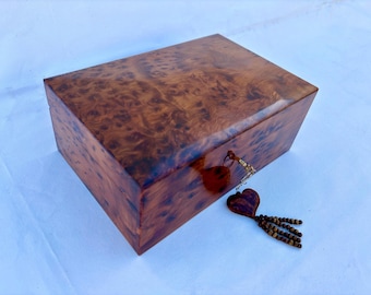 Puede incluir: Una caja de madera con un dije de llave en forma de corazón y un borla de cuentas. La caja está hecha de madera oscura con un patrón de veta natural. La caja tiene una cerradura y una llave.