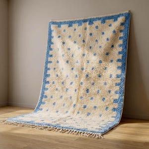 Puede incluir: Una alfombra rectangular con una base de color crema y un patrón repetitivo de pequeños puntos azules. La alfombra está enmarcada por un borde grueso azul con un diseño dentado e incluye flecos cortos de color claro en el borde inferior.
