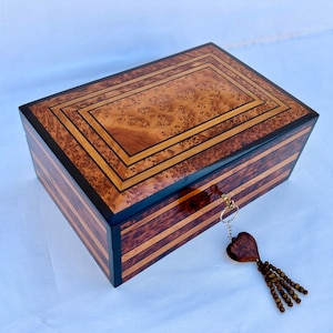 Puede incluir: Una caja de madera con una llave y un borla en forma de corazón. La caja está hecha de diferentes tipos de madera y tiene un borde negro. La caja está cerrada y la llave cuelga de la tapa.