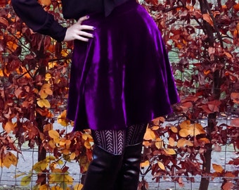May include: A woman wearing a black blouse with a bow tie and a purple velvet skirt. She is standing in front of a hedge with autumn foliage.