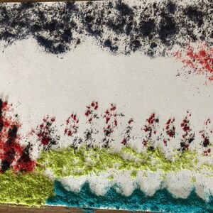 May include: A white background with a blue, red, and green sand art design. The design features a row of trees with red and black sand, a green grassy area, and a blue wavy line representing water.