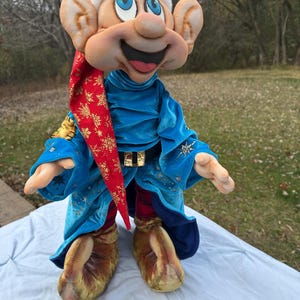 Puede incluir: Muñeco de peluche de un personaje de dibujos animados con orejas grandes, un gorro rojo y dorado y una túnica de terciopelo azul. El muñeco tiene una cara sonriente con mejillas rosadas y lleva un cinturón dorado. El muñeco está sobre una superficie blanca.