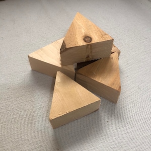 Może przedstawiać: Kilka niewykończonych drewnianych bloków w kształcie trójkąta ułożonych na jasnej, teksturowanej powierzchni. Bloki mają jasnobrązowy kolor, z widocznym usłojeniem i sękami. Bloki są ścięte pod kątem 45 stopni.