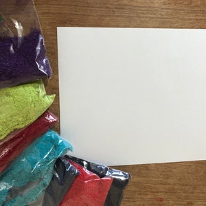 May include: Six small bags of colourful sand, arranged in a rainbow pattern, with a white sheet of paper to the right. The bags are clear plastic and contain purple, green, red, blue, black, and red sand.
