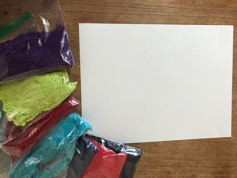 May include: Six small bags of colourful sand, arranged in a rainbow pattern, with a white sheet of paper to the right. The bags are clear plastic and contain purple, green, red, blue, black, and red sand.