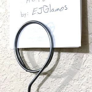 May include: A silver-toned metal sign holder featuring a circular wire loop at the top designed to grip a white rectangular card. The card displays the handwritten text Sign Holders by: EJGlamos in black ink against a plain white background.