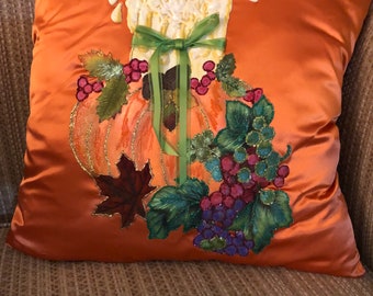 Puede incluir: Cojín decorativo de satén naranja con un diseño otoñal bordado. El diseño incluye una vela grande, calabazas, uvas y hojas de otoño. La vela tiene una cinta verde.