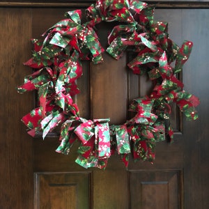Puede incluir: Una corona de Navidad de tela roja y verde con patrones de acebo y bayas. La corona está colgada en una puerta de madera marrón oscuro.