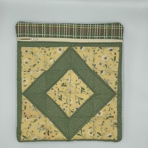 May include: A square quilted zippered pouch featuring a patchwork design with a central diamond motif. The fabric displays a yellow background with white daisy floral patterns and green fabric with white dots, finished with a green plaid top border.