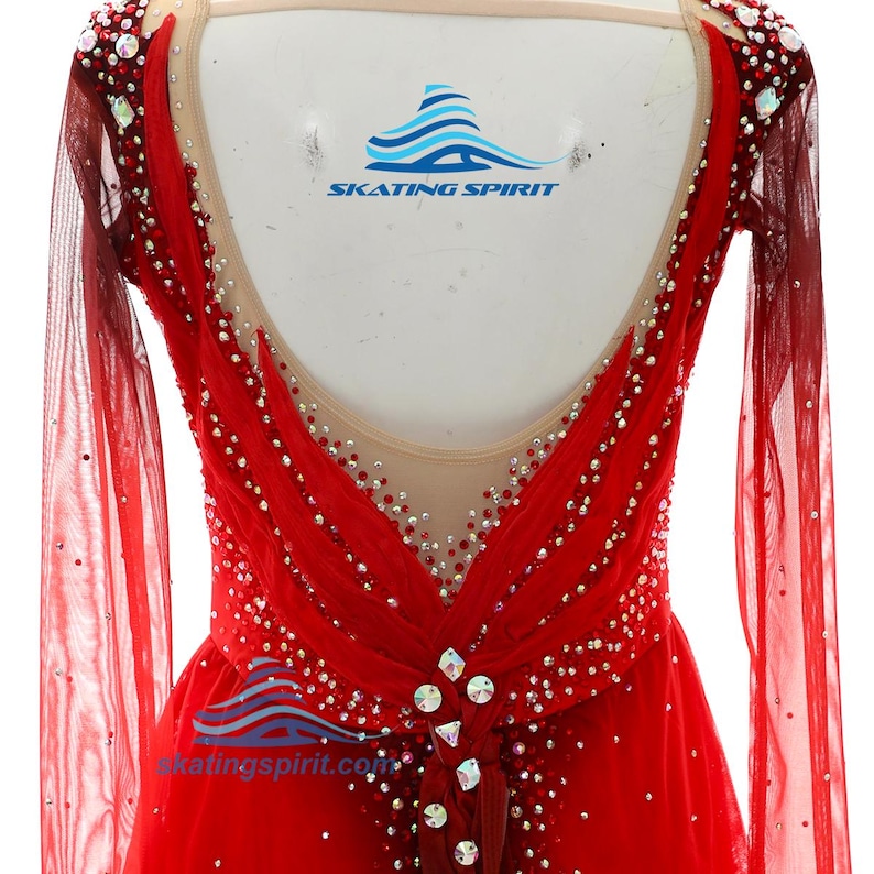 Peut inclure: Une robe de patinage artistique rouge &agrave; manches longues avec une encolure d&eacute;gag&eacute;e en maille nude, orn&eacute;e de nombreux cristaux rouges et transparents. Le v&ecirc;tement pr&eacute;sente des manches rouges transparentes et un n&oelig;ud central avec des cristaux.