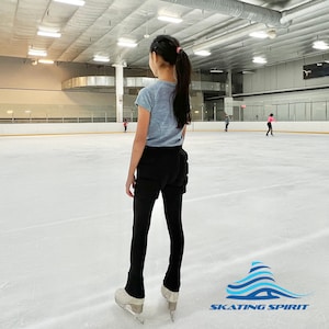 May include: A young person wearing black trousers and a light blue shirt is ice skating on a rink. The person is looking ahead and has their back to the camera. The rink is empty except for a few other people in the background. The text "SKATING SPIRIT" is visible in the lower right corner of the image.