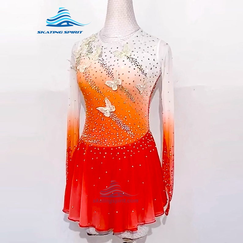 May include: A white and orange figure skating dress with a gradient design and butterfly embellishments. The dress has long sleeves and a short skirt. The skirt is red with a white and orange ombre effect. The dress is decorated with rhinestones and white butterflies.