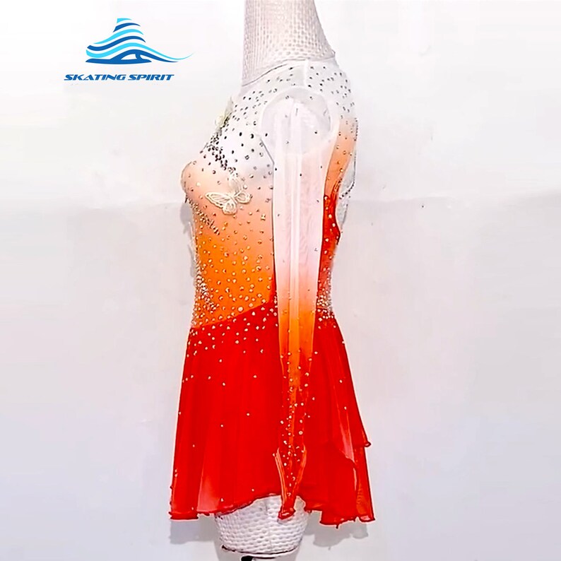 May include: A red, orange, and white figure skating dress with a butterfly design and rhinestones. The dress has long sleeves and a skirt with a layered design. "SKATING SPIRIT" is printed on the dress.