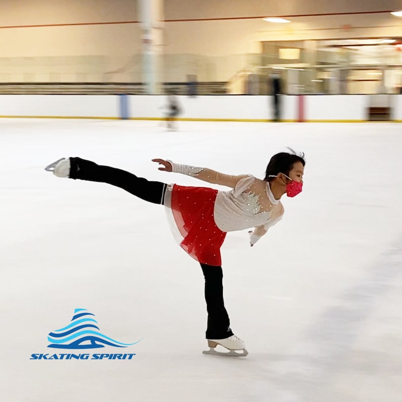 K&ouml;nnte beinhalten: Eine junge Eiskunstl&auml;uferin in einem wei&szlig;en Oberteil mit Pailletten, einem roten Rock und einer schwarzen Hose f&uuml;hrt eine Skaterbewegung auf einer Eisbahn aus. Die L&auml;uferin tr&auml;gt eine rosa Gesichtsmaske. Das Bild enth&auml;lt den Text "SKATING SPIRIT" in Blau und Wei&szlig;.