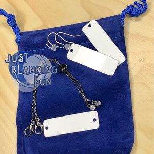 May include: A blue velvet pouch with a drawstring closure containing two blank rectangular metal tags, a silver chain necklace with two black beads, and a pair of silver earrings with clear beads. The pouch has the text "Just Blanking Fun" printed on it.