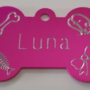 May include: Pink bone-shaped dog tag with silver engraved details. The tag has the name "Luna" engraved on it, along with a bone, fish, and bow.