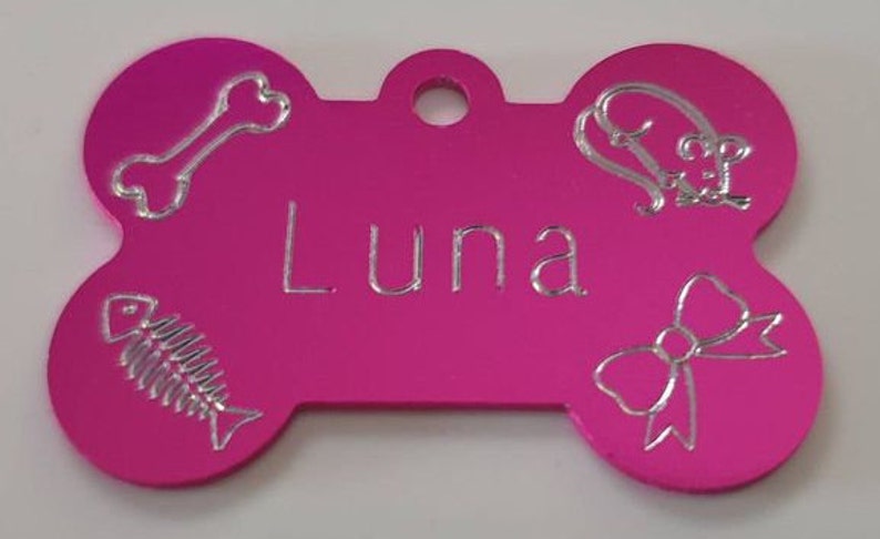 May include: Pink bone-shaped dog tag with silver engraved details. The tag has the name "Luna" engraved on it, along with a bone, fish, and bow.