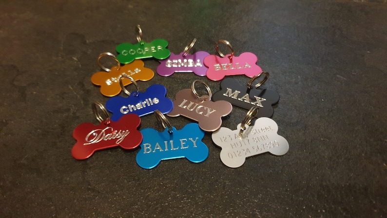 May include: A collection of ten colorful bone-shaped dog tags with engraved names, including Bailey, Lucy, Max, Charlie, Daisy, Bella, Gemma, Cooper, and a tag with a phone number.