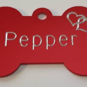 May include: Red bone-shaped dog tag with a silver inscription that reads "Pepper" and two silver hearts.