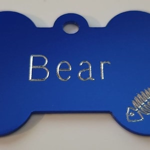 May include: Blue bone-shaped dog tag with the name "Bear" engraved in silver lettering and a silver fishbone design.