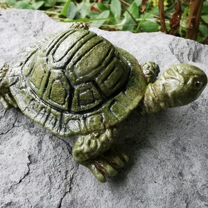 Pode incluir: Uma figura de tartaruga de cerâmica verde com uma casca detalhada e uma superfície texturizada. A tartaruga está sentada em uma pedra cinza.