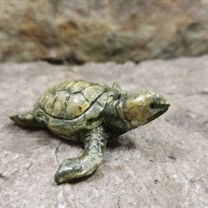 Pode incluir: Uma pequena figura de tartaruga de cerâmica verde e marrom. A tartaruga está em posição de caminhar com a cabeça virada para o lado.