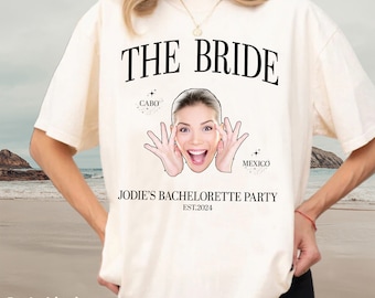 Könnte beinhalten: Weißes T-Shirt mit dem Text "The Bride" in Schwarz. Das Shirt zeigt außerdem das Gesicht einer Frau mit dem Text "Cabo" und "Mexico" über ihrem Kopf. Der Text "Jodie's Bachelorette Party Est. 2024" befindet sich unter dem Gesicht der Frau.