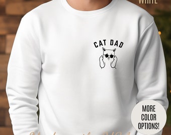 Może przedstawiać: Biała bluza z czarnym kotem w okularach przeciwsłonecznych, który pokazuje środkowy palec. Tekst "CAT DAD" znajduje się nad kotem. Tekst "Made in the USA" znajduje się na dole obrazu.