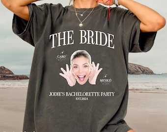 Könnte beinhalten: Ein graues T-Shirt mit dem Text "THE BRIDE" in Weiß. Das Shirt zeigt außerdem das Gesicht einer Frau mit dem Text "CABO" und "MEXICO" über ihrem Kopf. Der Text "JODIE'S BACHELORETTE PARTY EST.2024" befindet sich unter dem Gesicht der Frau.