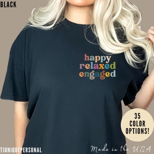 以下が含まれることがあります： カラフルな文字で「happy relaxed engaged」と書かれた黒いTシャツ。テキストは縦に積み重ねられ、シャツの中央に配置されています。シャツは、長いブロンドの髪をした人が着用しています。画像の右下隅には「35 COLOR OPTIONS！」というテキストが表示されています。画像の右下隅には「Made in the USA」というテキストが表示されています。