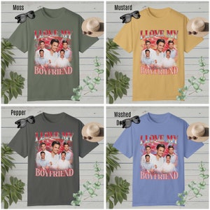 May include: Four different color t-shirts with a graphic print of a group of men with the text "I Love My Boyfriend" in red and white letters. The shirts are displayed on a white wooden background with plants and sunglasses.