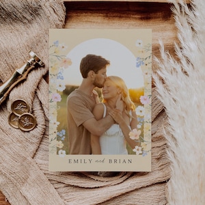 May include: A wedding invitation featuring a couple embracing, framed by a floral arch. The invitation has the names "EMILY and BRIAN" at the bottom. The background is a warm, neutral tone, with a soft, romantic aesthetic.