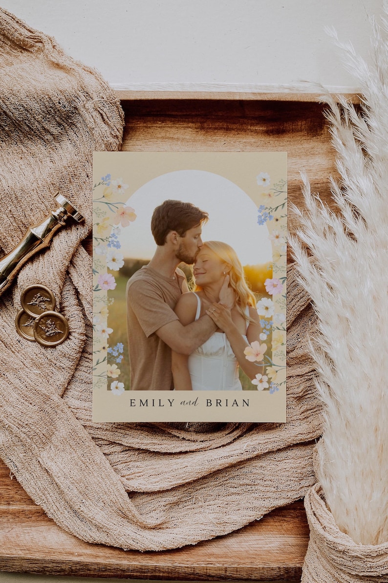 May include: A wedding invitation featuring a couple embracing, framed by a floral arch. The invitation has the names "EMILY and BRIAN" at the bottom. The background is a warm, neutral tone, with a soft, romantic aesthetic.