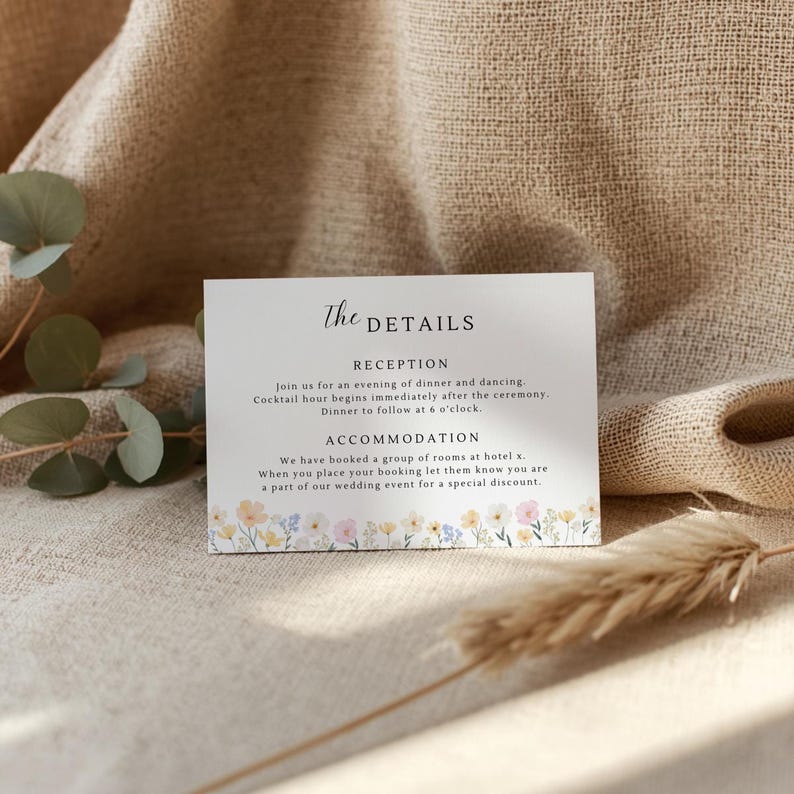 May include: A white card with the text "The Details" and wedding reception information. The card has a floral border with yellow, pink, and blue flowers. The card is on a beige linen surface.