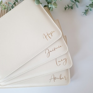 May include: A stack of four off-white, rectangular pouches with gold lettering. The names "Hope", "Jaimie", "Lucy", and "Amber" are written on the front of each pouch. The pouches are arranged in a fan shape.