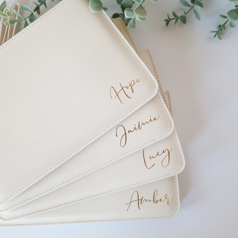 May include: A stack of four off-white, rectangular pouches with gold lettering. The names "Hope", "Jaimie", "Lucy", and "Amber" are written on the front of each pouch. The pouches are arranged in a fan shape.