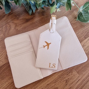 May include: A white passport holder with a matching luggage tag. The luggage tag has a gold airplane silhouette and the initials "LS" in gold.