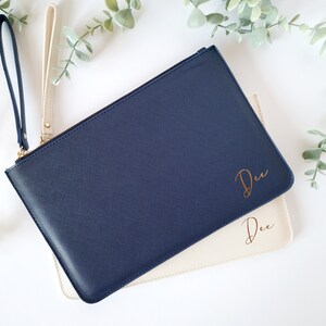 May include: Two leather wristlet clutches, one navy blue and one cream. The navy clutch has a gold zipper and a wrist strap. The word "Dee" is written in gold script on both clutches. Greenery is in the background.