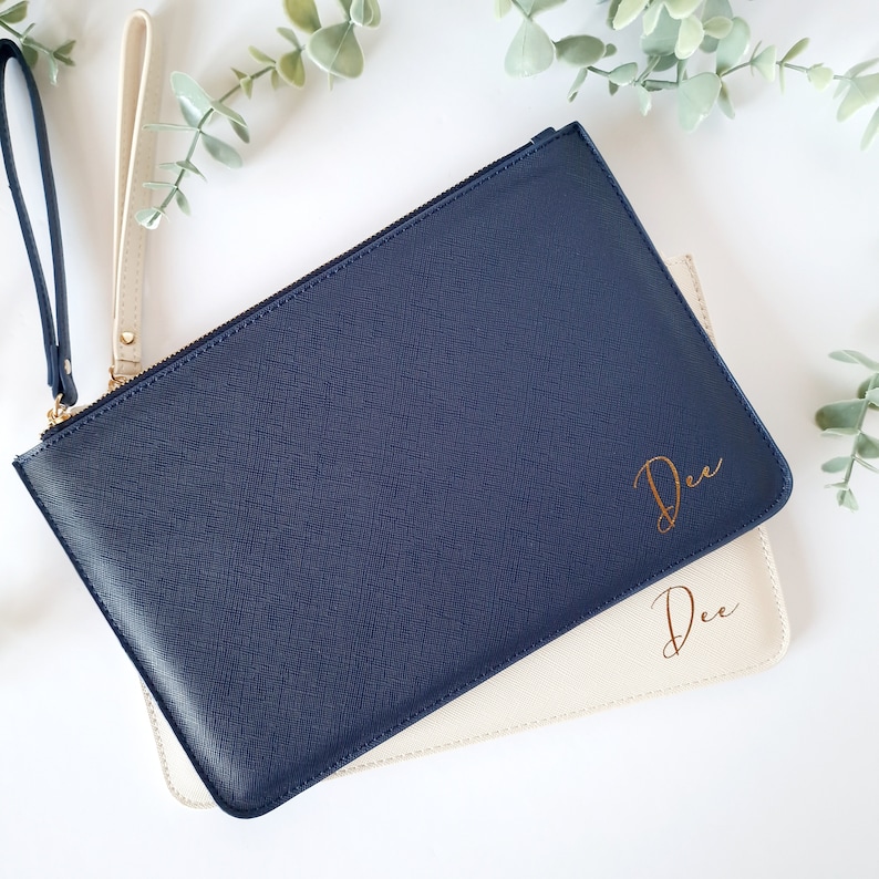 May include: Two leather wristlet clutches, one navy blue and one cream. The navy clutch has a gold zipper and a wrist strap. The word "Dee" is written in gold script on both clutches. Greenery is in the background.