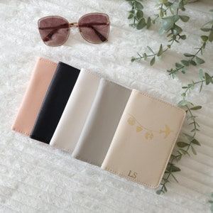 May include: Five passport holders in different colors, including beige, black, white, gray, and a light beige with a gold airplane and hearts design. The light beige passport holder has the initials "LS" in gold.