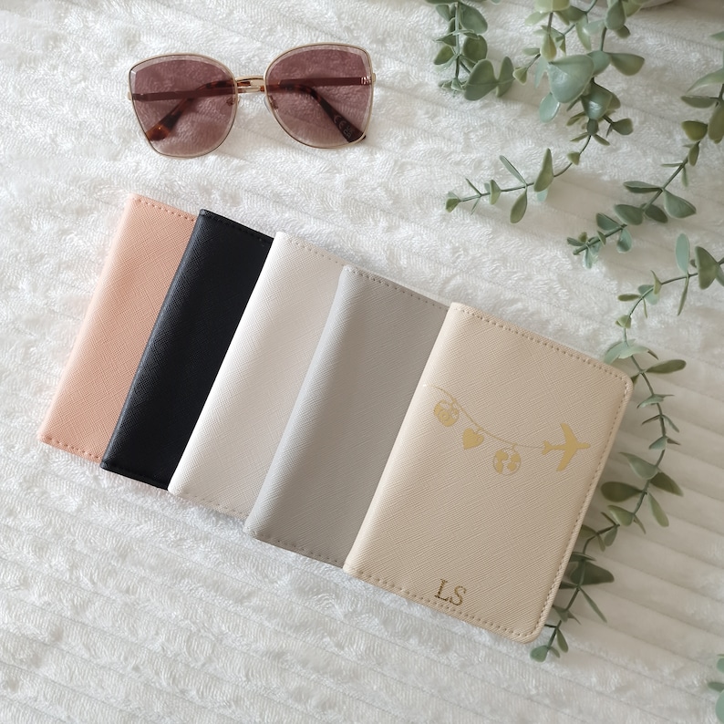 May include: Five passport holders in different colors, including beige, black, white, gray, and a light beige with a gold airplane and hearts design. The light beige passport holder has the initials "LS" in gold.