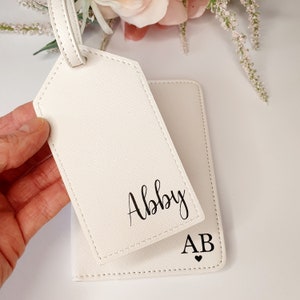 May include: White leather luggage tag with the name Abby written in black cursive. A white passport holder with the initials AB and a small heart.