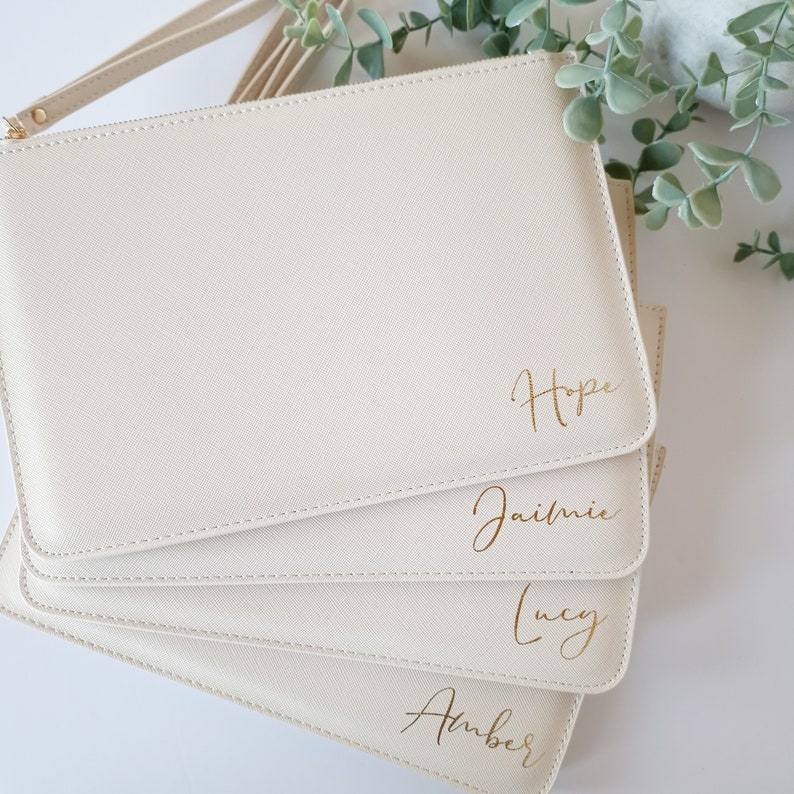 May include: Cream-colored clutch purses with gold lettering. The purses are stacked, with the names "Hope", "Jaimie", "Lucy", and "Amber" visible. Each purse has a wrist strap and a gold zipper.